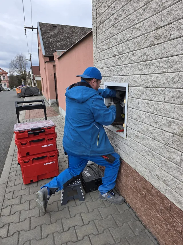 Montér v&nbsp;modré pracovní bundě opravuje plynoměr ve skříni na fasádě domu