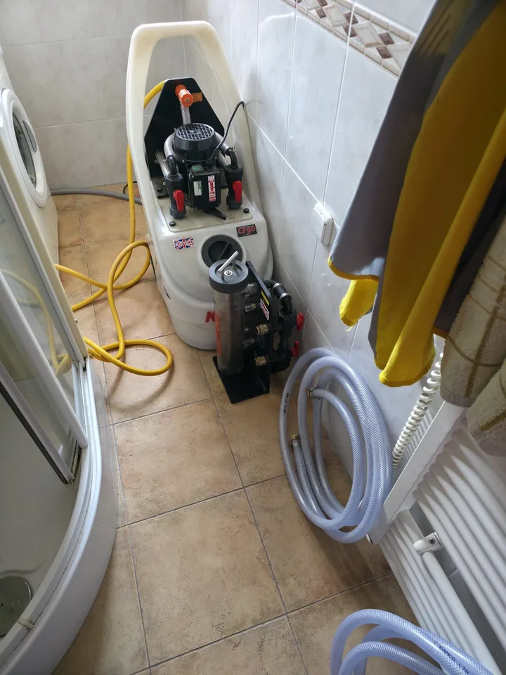 Proplachovací stroj s filtrem a smotanými hadicemi postavený na dlažbě v koupelně u sprchového koutu